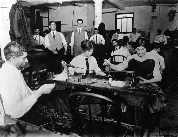 Workers At A Small Bench Hand Finish Garments While Managers Look On